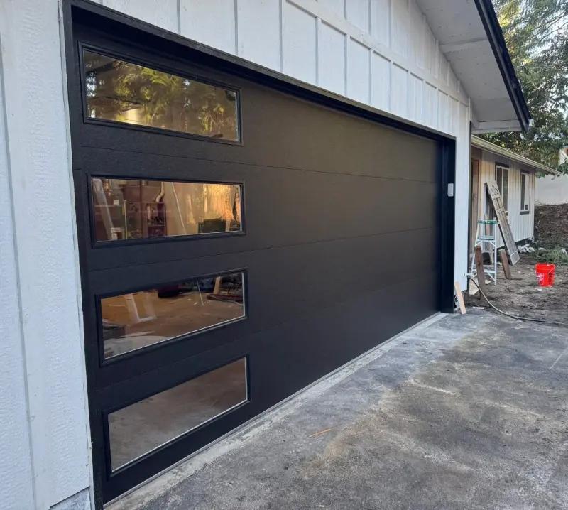 A black modern steel garage door with a column of glass lites on a white board-and-batten house.