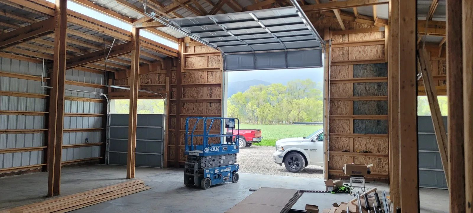 Inside a new steel shop under construction, with a wide overhead door open onto green fields.